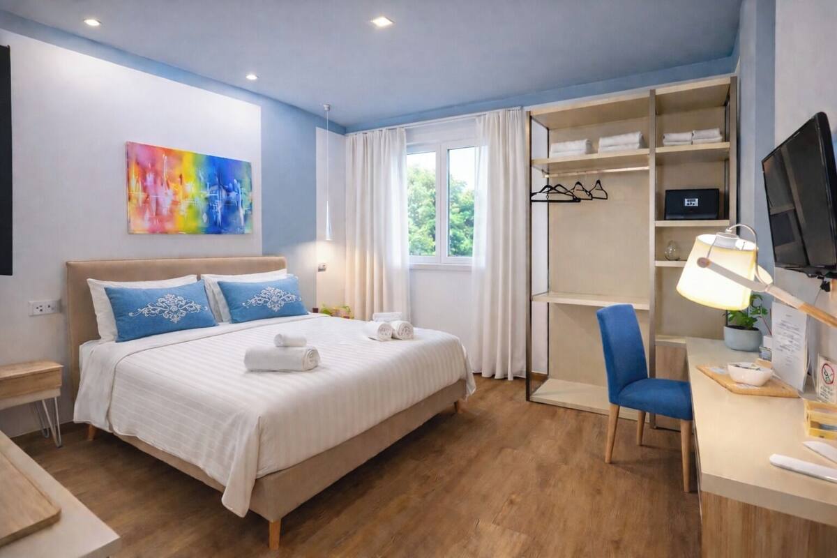 Habitación de hotel con cama, escritorio, silla, televisor, ventana con cortinas y un cuadro de colores en la pared.