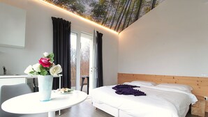 Quarto de hotel moderno com uma cama, uma mesinha e uma cadeira. Há uma janela com cortinas, e um vaso com flores na mesa.