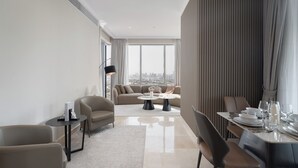 Ein modernes Hotelzimmer mit großem Fenster und Blick auf die Stadt, einer Couch, einem Esstisch für zwei Personen und einem kleinen Beistelltisch.
