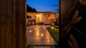 Een houten poort met libellenornamenten, die leidt naar een huis met een verharde oprit en buitenverlichting.