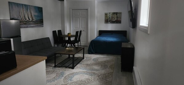 Quarto compacto com cama, sofá, mesinha e televisão.