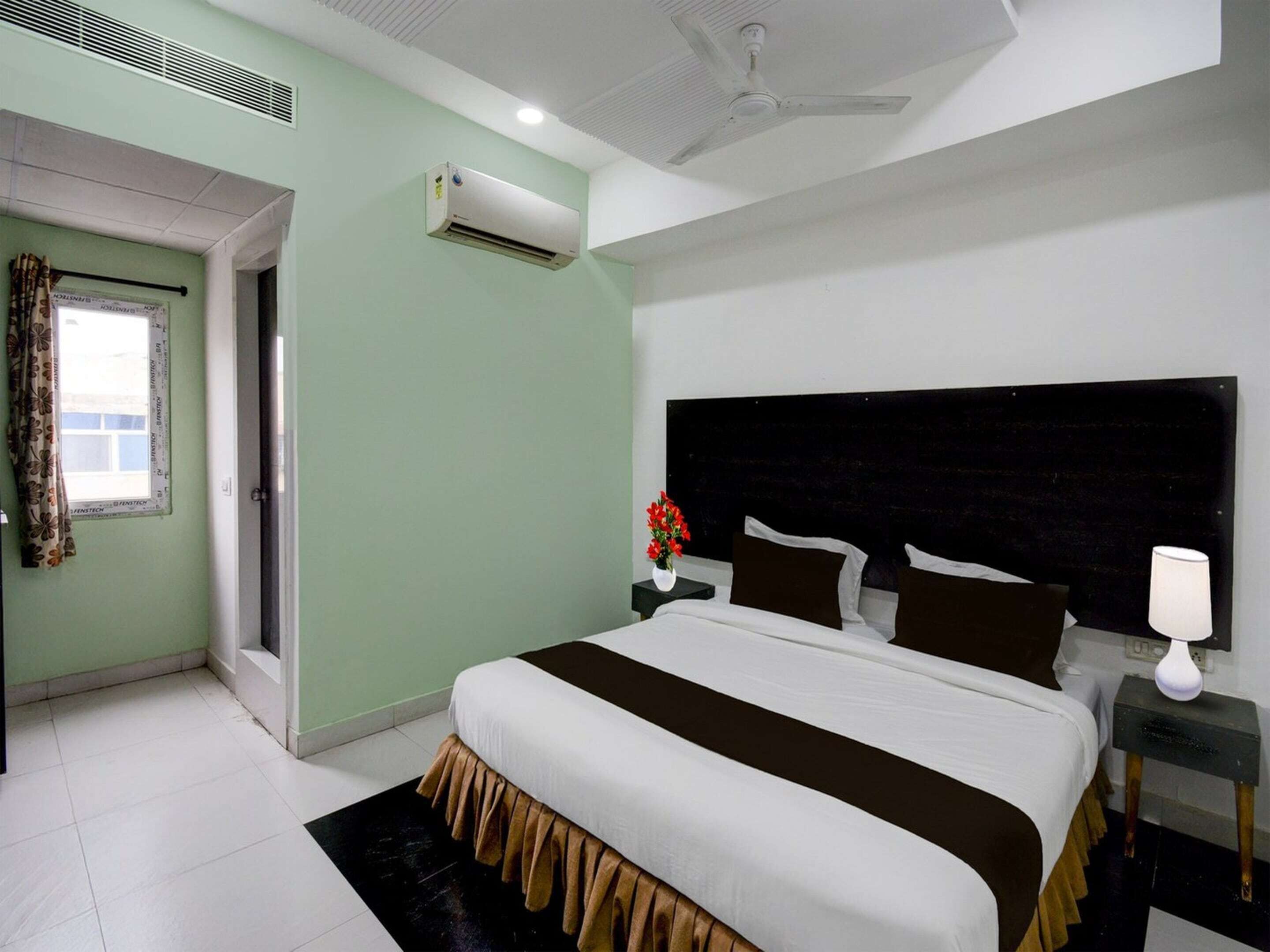 A hotel room with a bed, a nightstand, a lamp, an air conditioning unit, and a window with curtains.