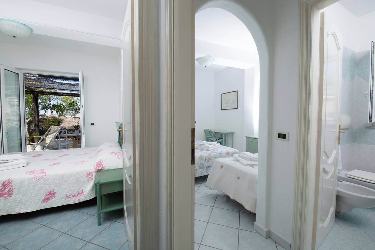 A bedroom with a bed, a desk, and a chair. There is a bathroom with a toilet and a shower. The room has white walls and a tiled floor.