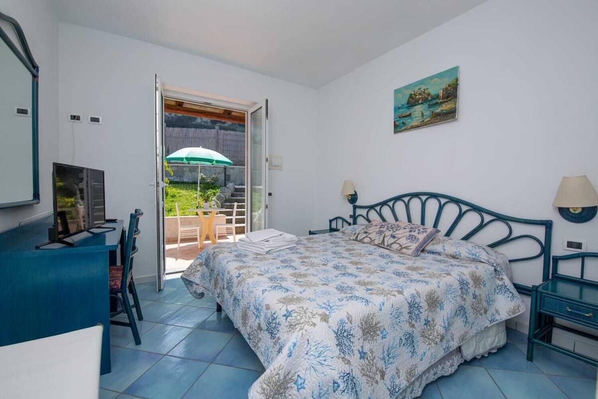 A bedroom with a bed, a desk, and a chair. There is a window with a view of a garden and a painting on the wall.