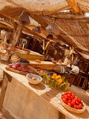 A rustic wooden counter with various fruits and vegetables, a woven thatched roof, and a wooden interior.