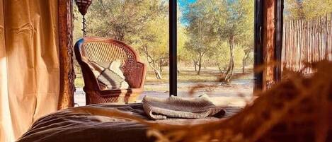 A room with a bed, a chair, and a view of trees through large windows.