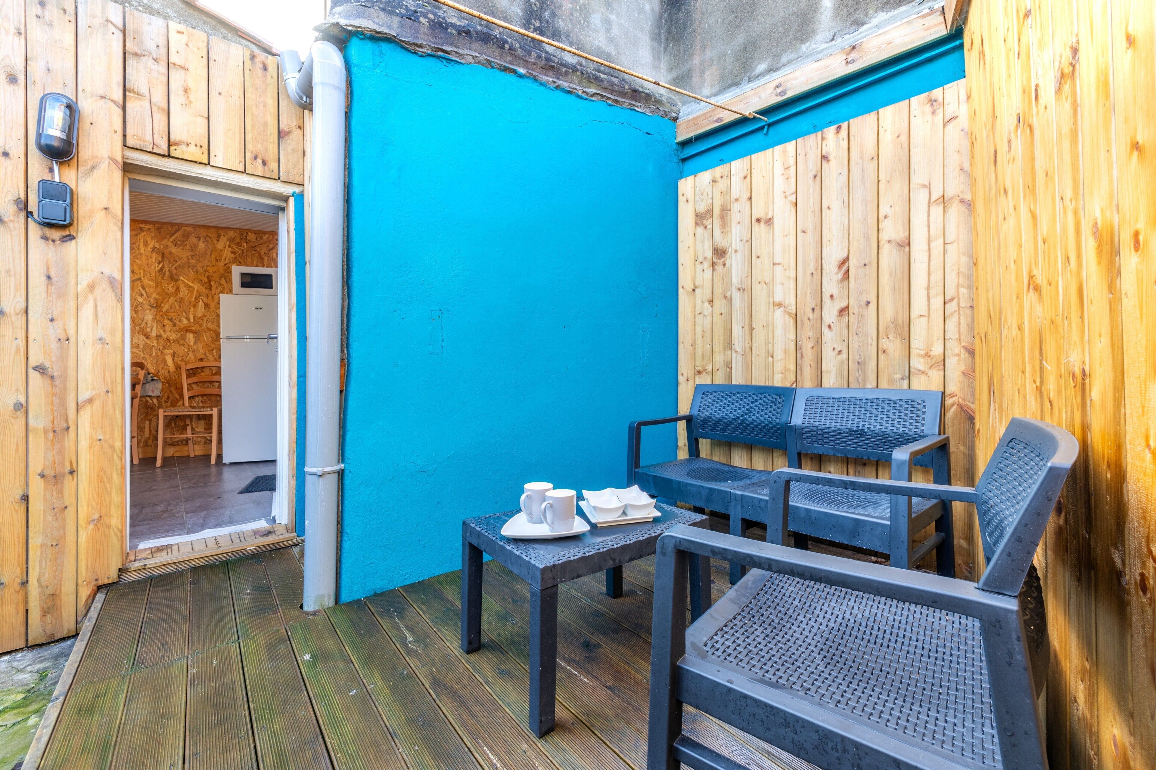 Una terraza de madera con un muro azul, un refrigerador blanco y una mesa con sillas.