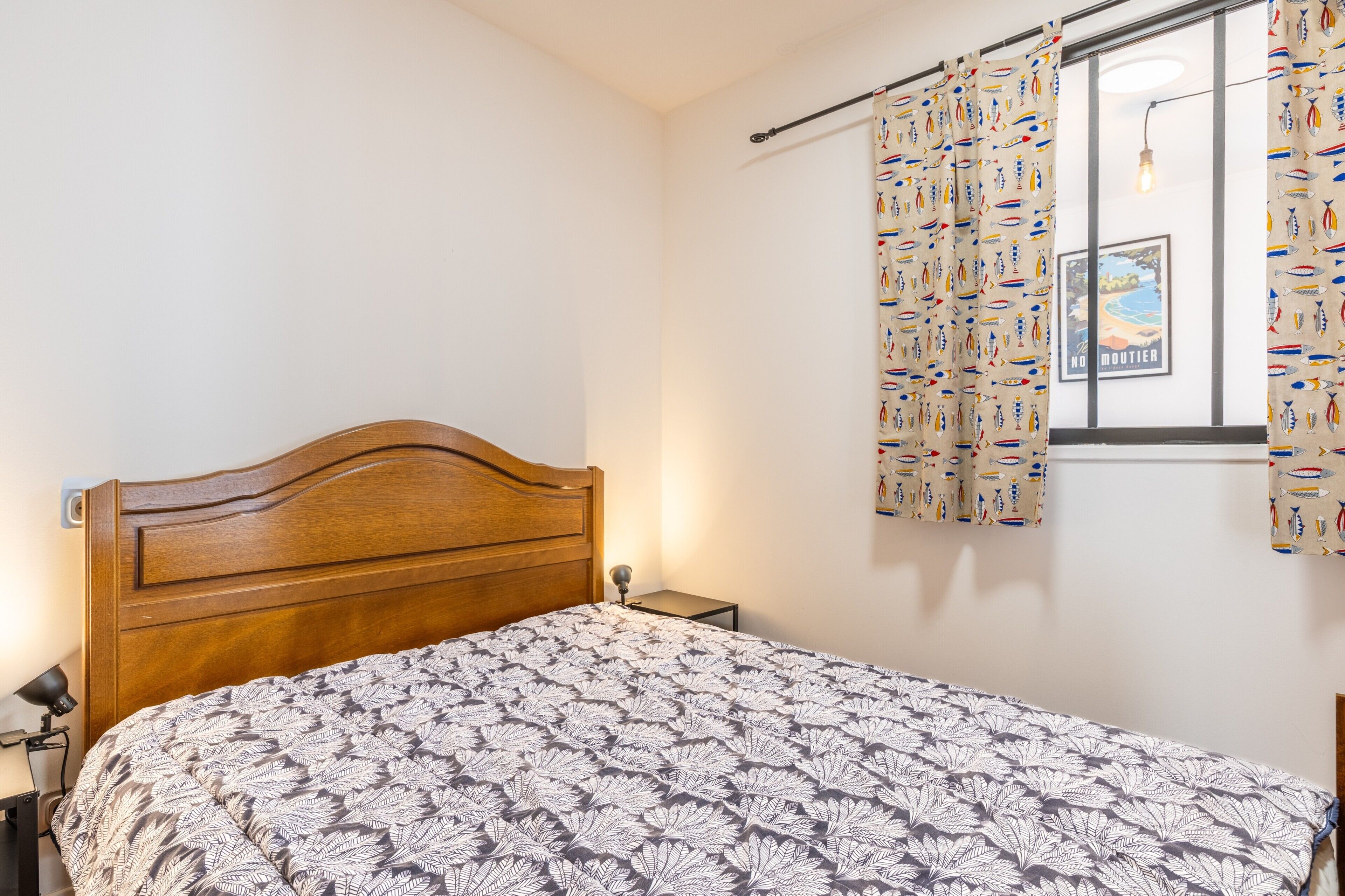 A bedroom with a wooden headboard, a patterned bedspread, a nightstand, a lamp, and a window with curtains.