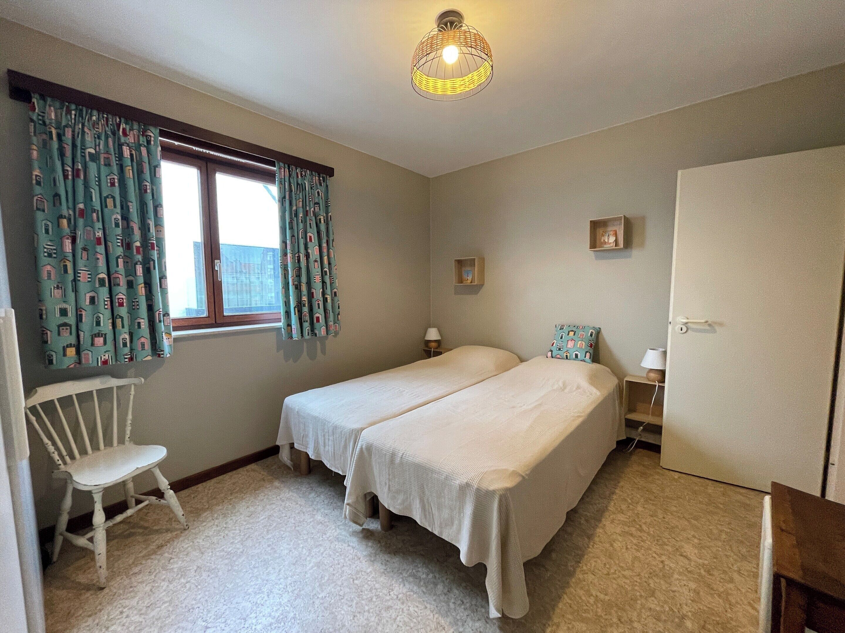 A bedroom with two single beds, a window with curtains, a chair, bedside tables, and a door.