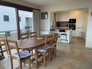 A modern kitchen with white cabinets and stainless steel appliances, a dining area with wooden chairs and a table, and a balcony with large windows and curtains. - Seaside Apartment by the Zeedijk Promenade (Koksijde)
