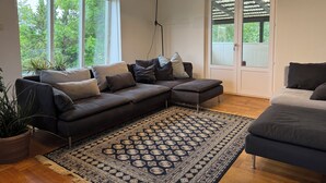 Un salon moderne avec un canapé d’angle, un tapis à motifs et de grandes fenêtres donnant sur un paysage verdoyant.