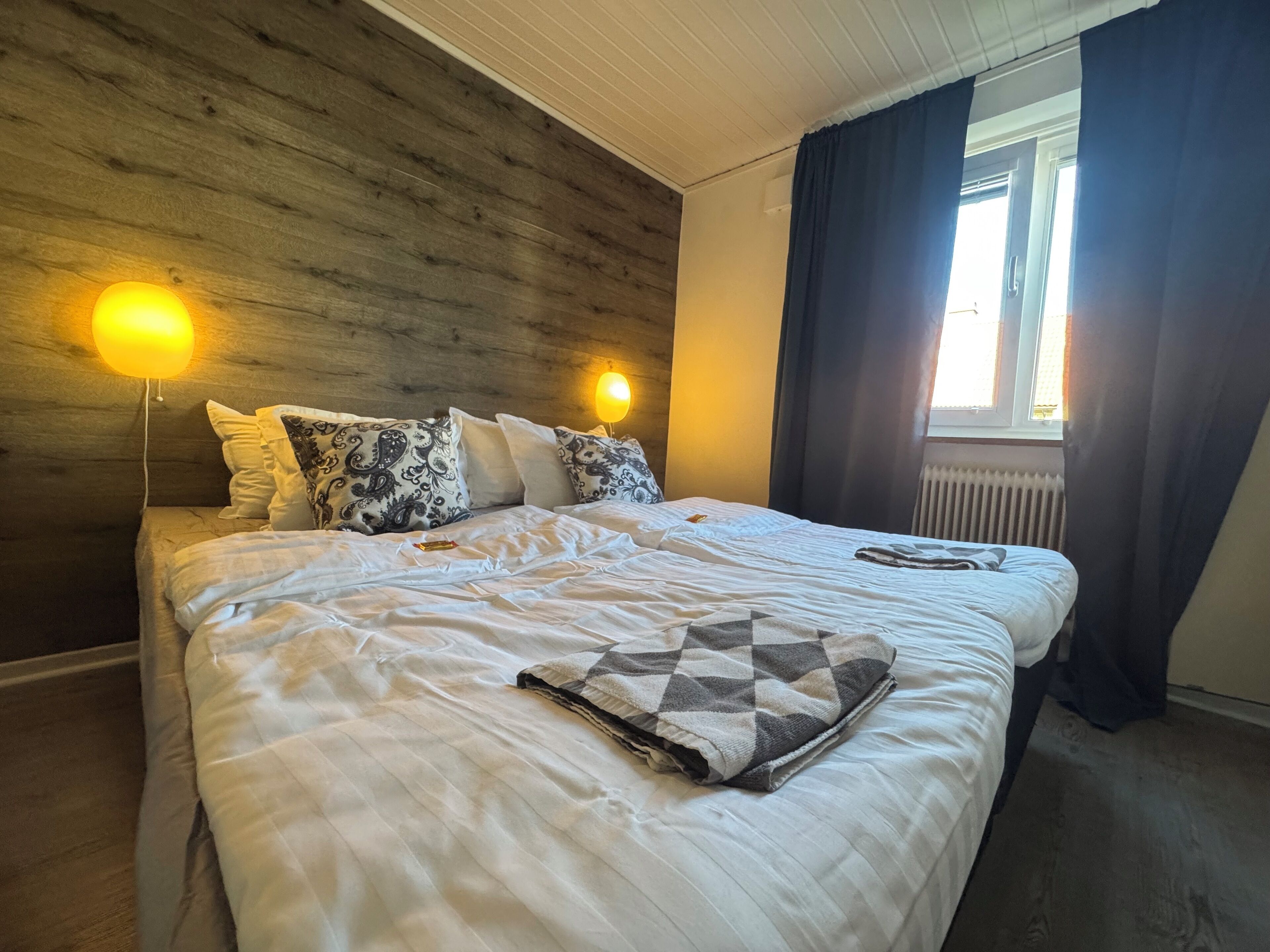 Una cama bien tendida con un edredón estampado, dos lámparas de noche y una ventana con cortinas.