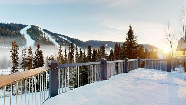 A snow-covered deck with a metal railing, overlooking a forested mountain slope. - Ski-In Hot Tub | Sunburst Drive Mountain Home (Sun Peaks)