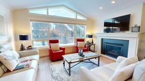 A spacious living room with a fireplace, large windows with a view of snow-covered trees, and modern furniture. - Ski-In Hot Tub | Sunburst Drive Mountain Home (Sun Peaks)