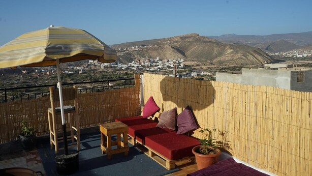 A terrace with a wooden table, two red cushioned benches, a striped umbrella, and a view of a town and mountains in the background. - titryte (Aourir)