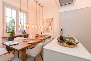A modern kitchen with a wooden dining table, white chairs, hanging pendant lights, and a fruit basket on the island. - MGH Luxury - Sunset Lakeview House Desenzano (Desenzano del Garda)