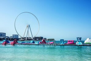 Ein Ufergebiet mit einem großen Riesenrad, bunten schwimmenden Plattformen mit Sitzgelegenheiten und freiem Blick auf die Skyline der Stadt im Hintergrund.