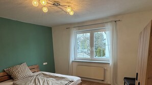 Ein Schlafzimmer mit einem hölzernen Bett, einem Fenster mit Vorhängen, einem Heizkörper und einer Deckenleuchte.