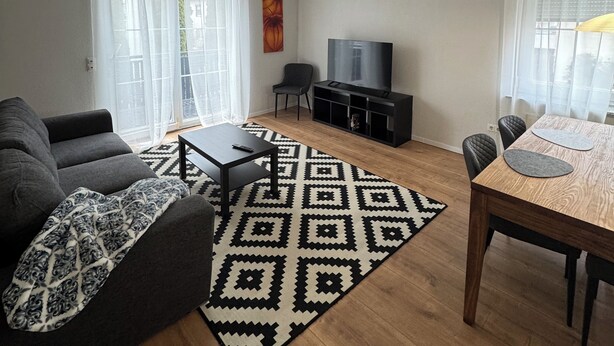 Ein Wohnzimmer mit einer Couch, einem Couchtisch, einem Fernseher, einem Esstisch mit Stühlen und einem gemusterten Teppich.