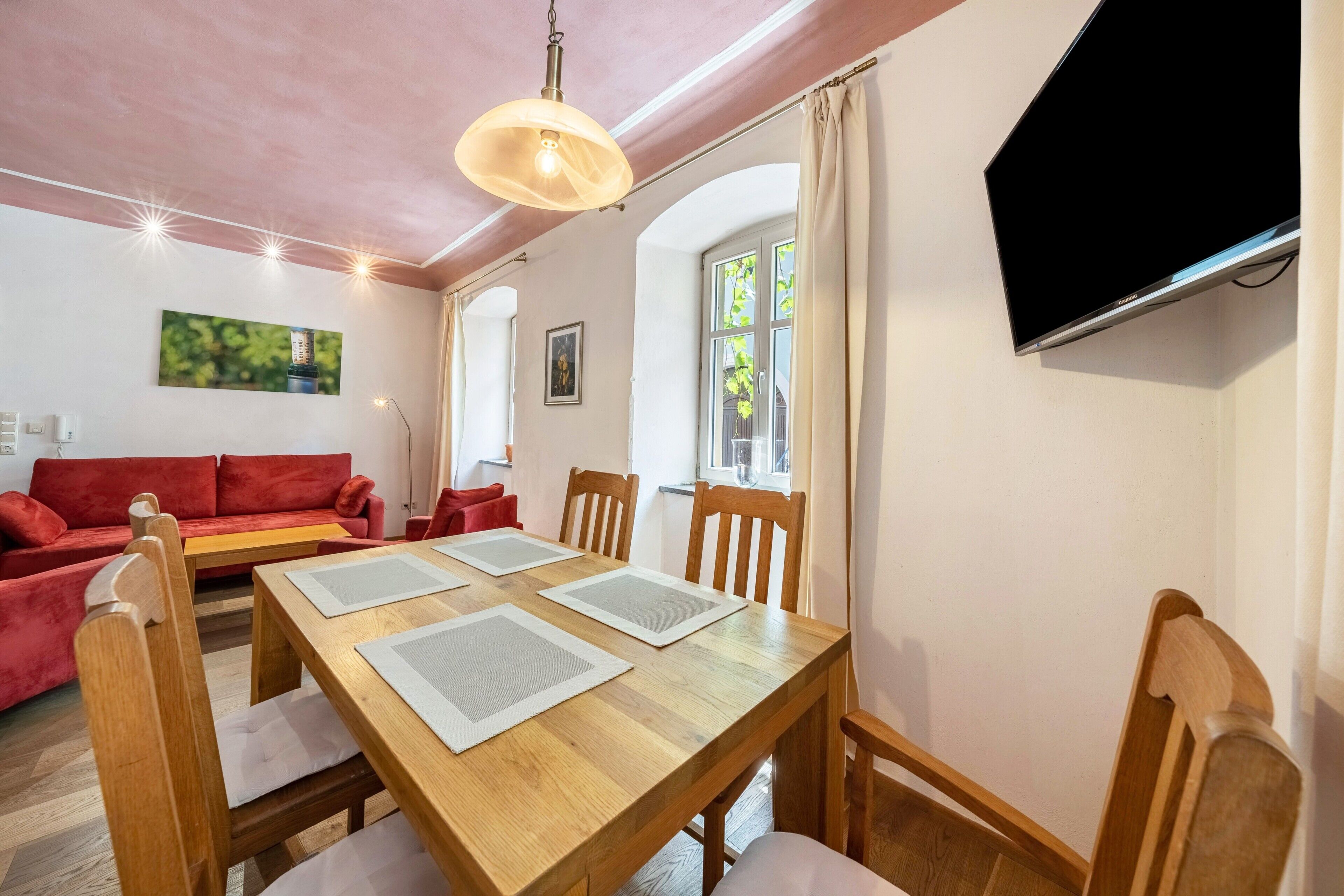 Ein Essbereich mit einem Holztisch, Stühlen, einer roten Couch und einem an der Wand montierten Fernseher.