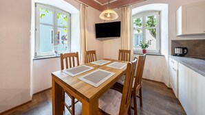 Een eetgedeelte met een houten tafel, stoelen, een aan de muur bevestigde televisie en ramen voorzien van gordijnen.