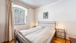 Een slaapkamer met een bed, een raam met gordijnen, een nachtkastje met een lamp en een aan de muur hangend schilderij.
