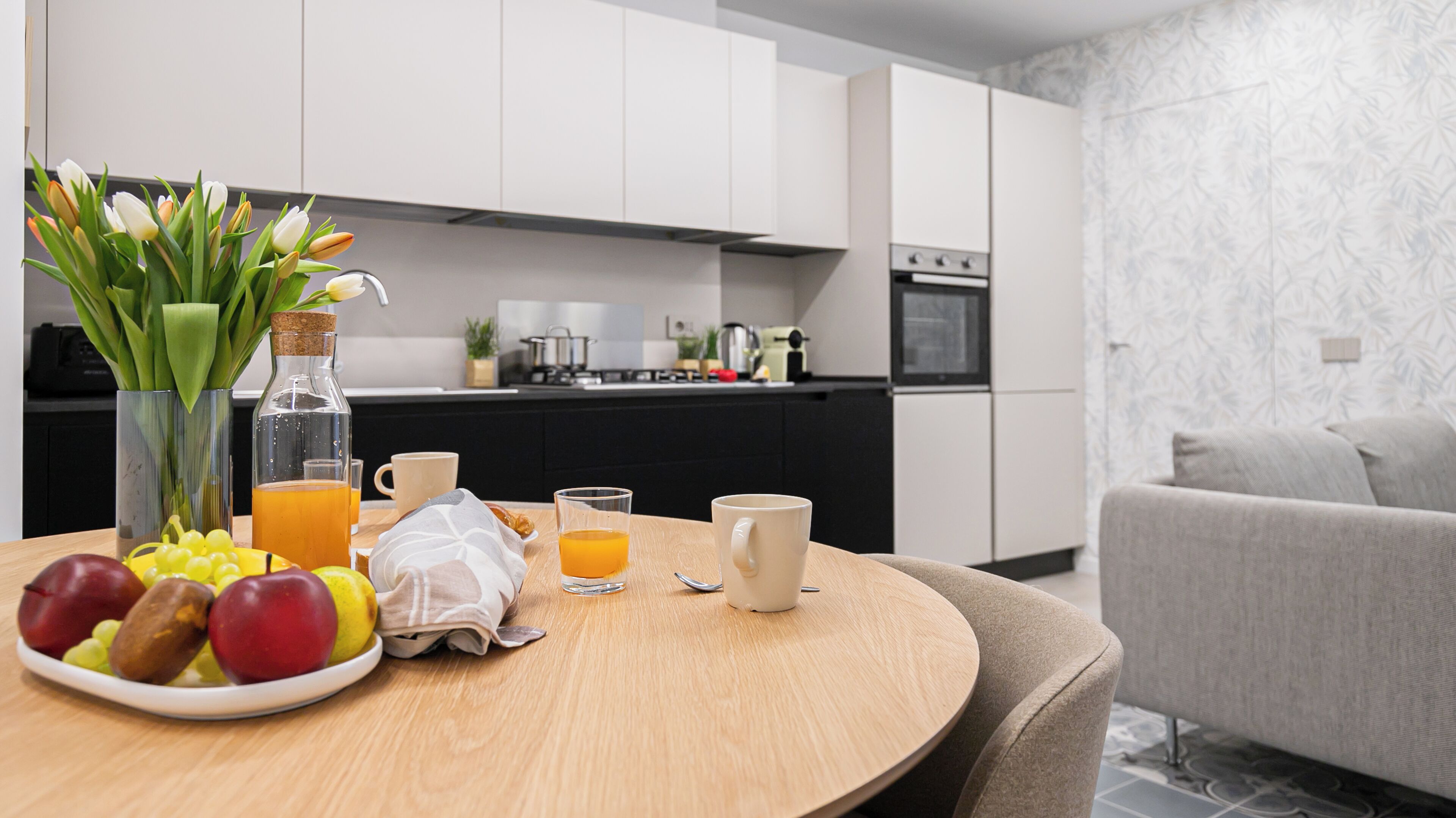 A modern kitchen with white cabinets, a dining table with fruits, glasses of orange juice, and a vase with flowers.