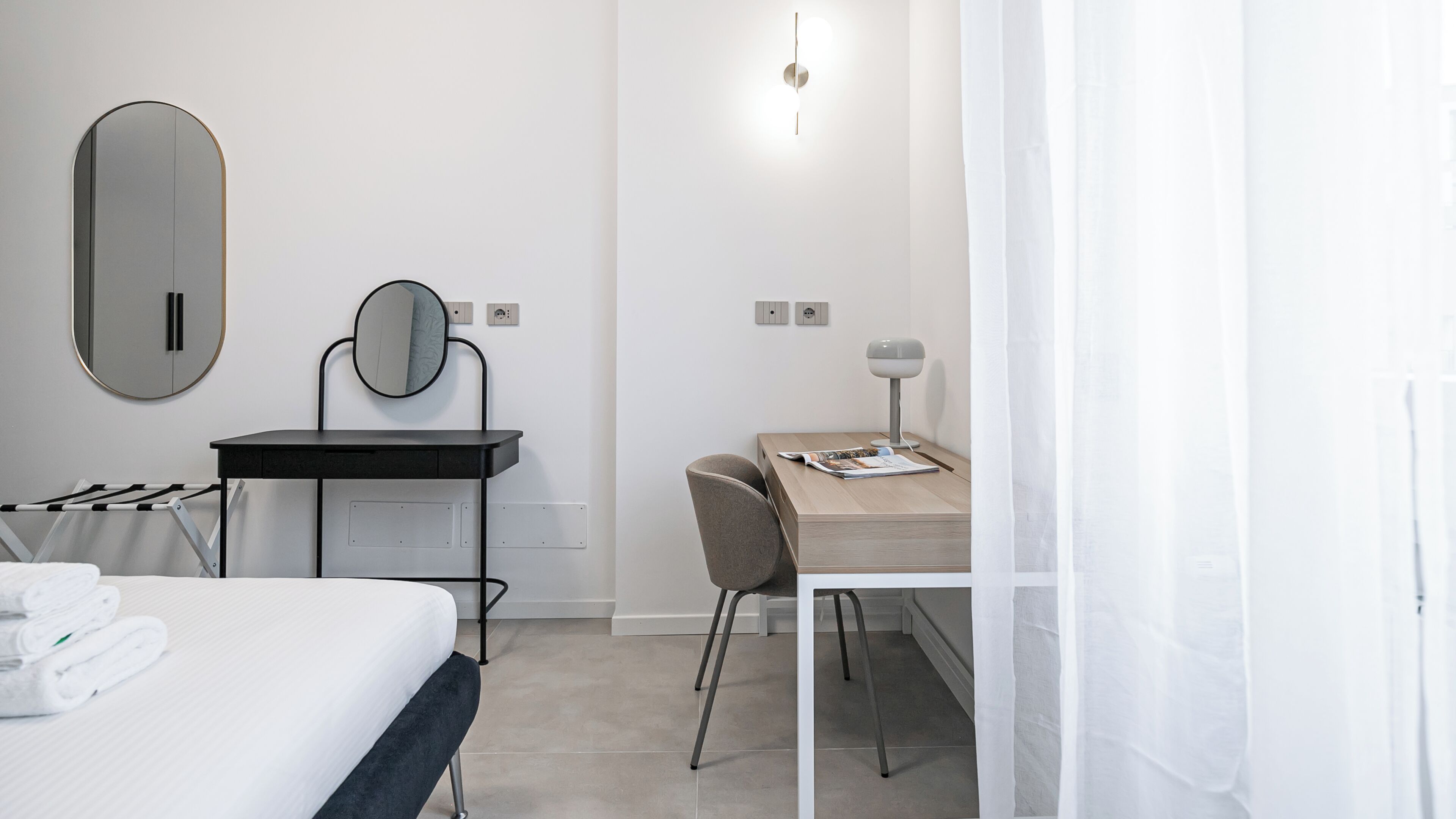 A modern bedroom with a bed, a desk, a chair, a mirror, and a bedside table.