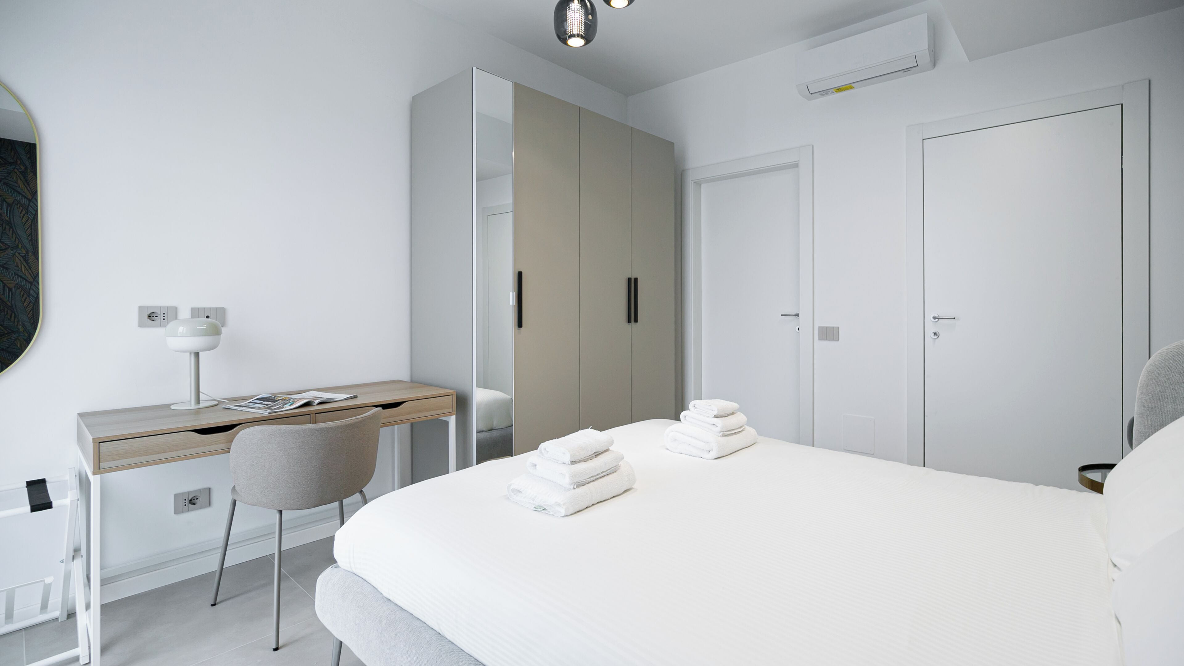 A modern hotel room with a bed, a desk, and a chair. There are two doors and an air conditioning unit on the wall.