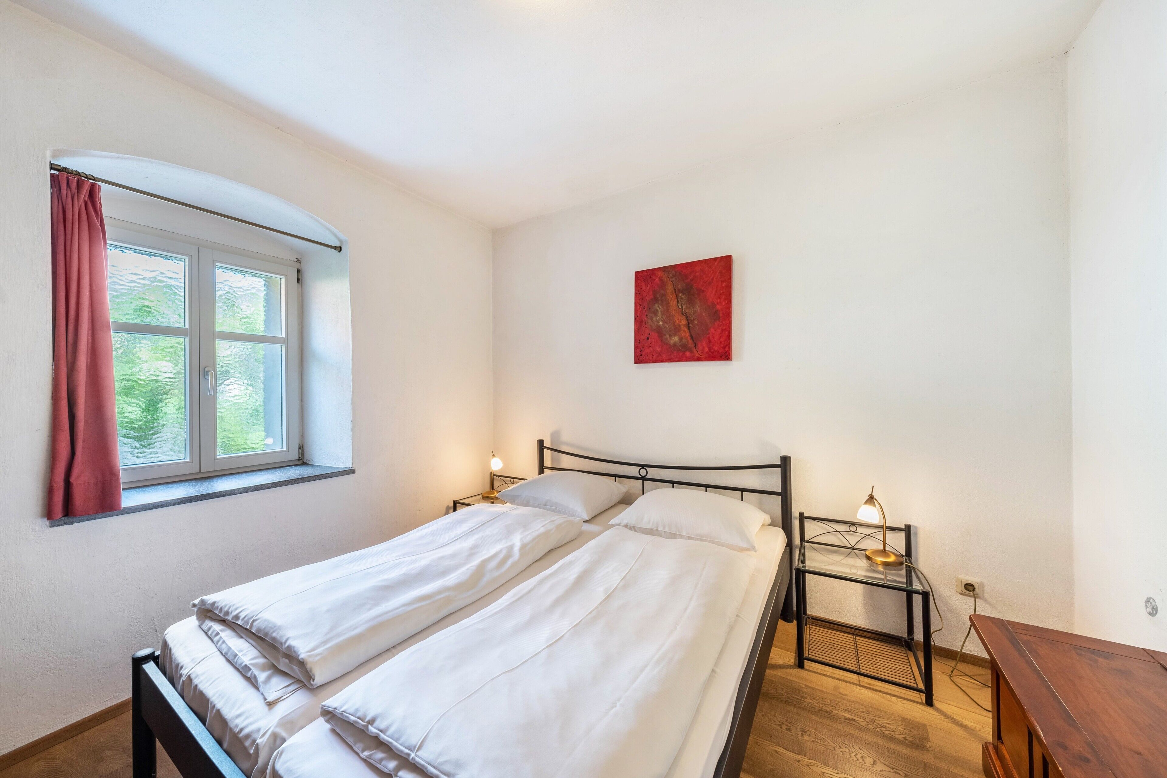 Une chambre avec deux lits, une fenêtre avec des rideaux, une table de chevet avec une lampe et une œuvre d’art rouge accrochée au mur.