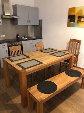 Una mesa de comedor de madera con bancos, una zona de cocina con armarios grises y electrodomésticos de acero inoxidable, y un cuadro en la pared.
