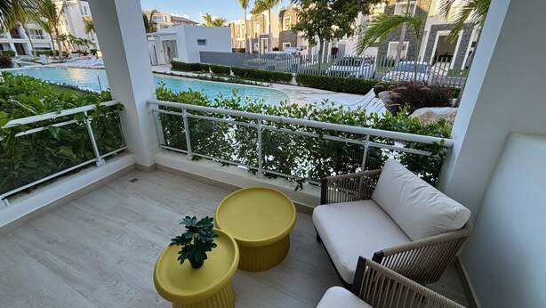 En balkon med udsigt over en swimmingpool, hvide møbler og runde, gule borde.