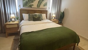 Et soveværelse med en stor seng, to sengeborde med lamper, en sengegavl med bladmotiv og en stor plante på den ene side.