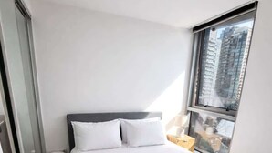 Ein modernes Hotelzimmer mit einem großen Bett, weißen Bettwäsche, einem dunklen Kopfteil und Blick auf eine Stadtkulisse durch ein großes Fenster.