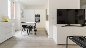Ett modernt kök med vita skåp, ett matbord med stolar, en inbyggd ugn och en platt-TV.