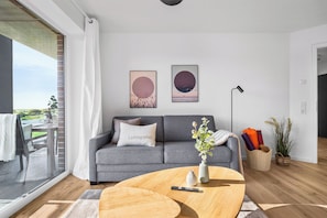 Ein modernes Wohnzimmer mit grauem Sofa, einem Couchtisch und einem großen Fenster mit Blick auf einen Balkon.