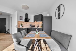 A modern kitchen with a dining area, featuring a wooden table, grey upholstered chairs, and a black-framed clock on the wall. - The View C05 Sonnengruß - Balcony, garden furniture, shared. Sauna, car park (Dagebüll)