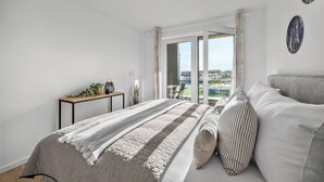 A bedroom with a bed, a nightstand, a window with curtains, and a view of the outside. - The View C05 Sonnengruß - Balcony, garden furniture, shared. Sauna, car park (Dagebüll)