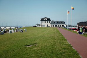A grassy area with a pathway leading to a white building with a black roof. There are flags on flagpoles to the right. - The View C05 Sonnengruß - Balcony, garden furniture, shared. Sauna, car park (Dagebüll)