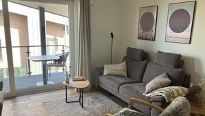 Una sala de estar con un sofá gris, una mesita redonda pequeña, un sillón mullido y un ventanal con vista a un edificio de ladrillo.