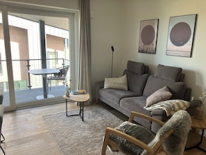 Ein Wohnzimmer mit grauem Sofa, einem kleinen runden Tisch, einem kuscheligen Sessel und einem großen Fenster mit Blick auf ein Ziegelgebäude.