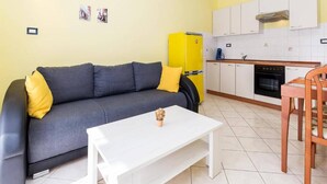 Un espacio habitable compacto con sofá, una mesita blanca, un refrigerador amarillo, una zona de cocina con armarios y estufa, y una silla de madera.