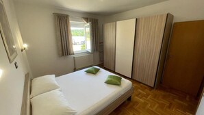Un dormitorio con cama, ventana con cortinas, radiador, armario y puerta.