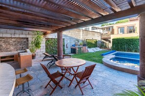 Eine Terrasse im Freien mit einem runden Holztisch und Stühlen, einem Steinkamin und einem Swimmingpool.