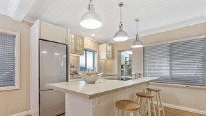 A modern kitchen with a stainless steel refrigerator, a large island with stools, pendant lighting, and a view of the adjacent dining area. - Metawisebnb Waterfront 3BR Coastal Escape (Deception Bay)