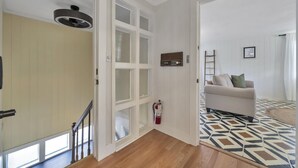 En hall med trägolv, en vit dörr, en vit vägg med en inramad tavla och en beige soffa med kuddar i bakgrunden.