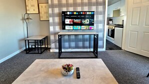 A living room with a flat-screen TV mounted on a wall, a small table with a plant, and a remote control on a coffee table. - Bright and Cozy Home Near Hampden Park (Glasgow)