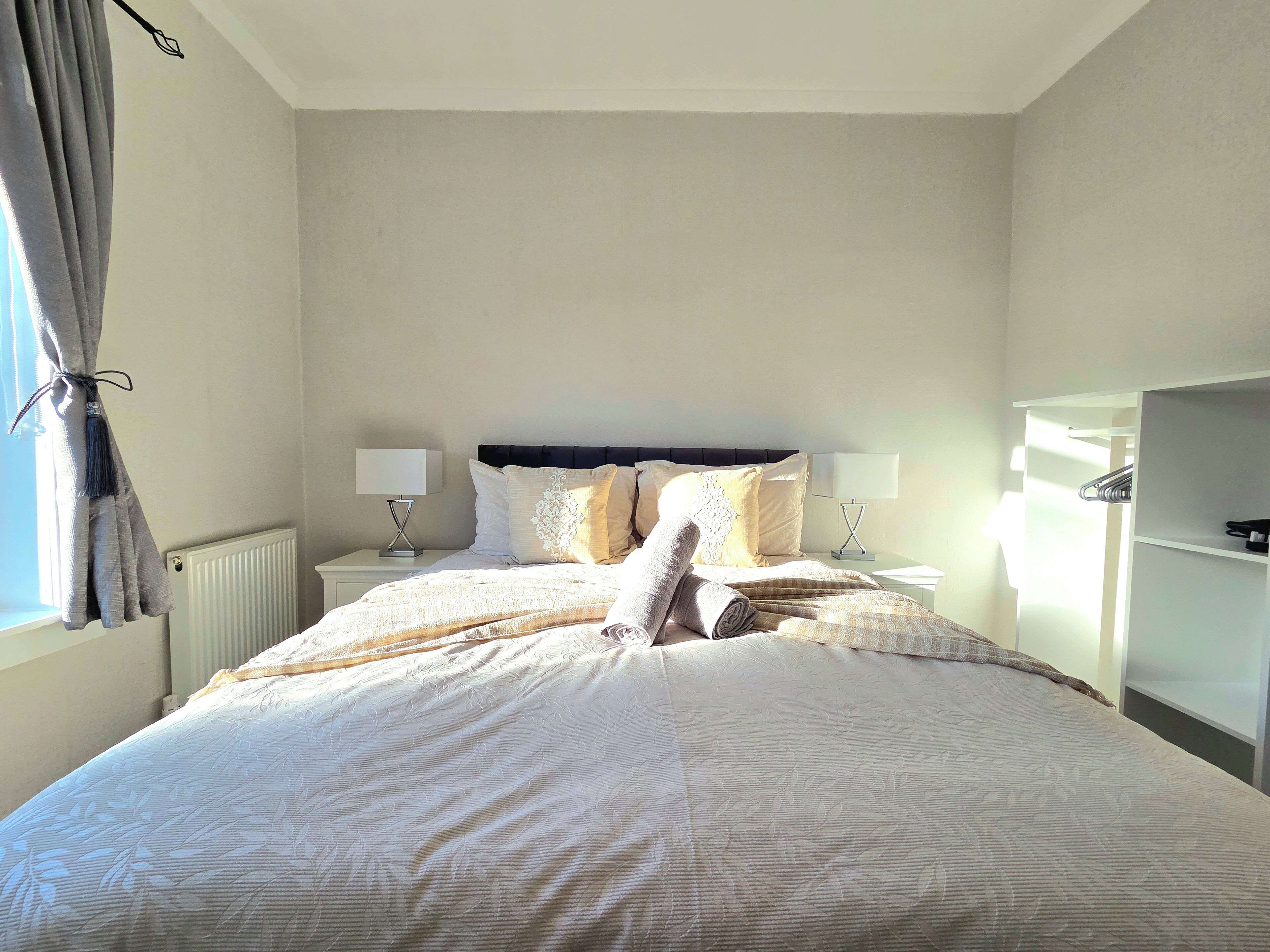 Un lit bien fait, avec une tête de lit, deux tables de chevet avec des lampes, un radiateur et une armoire.