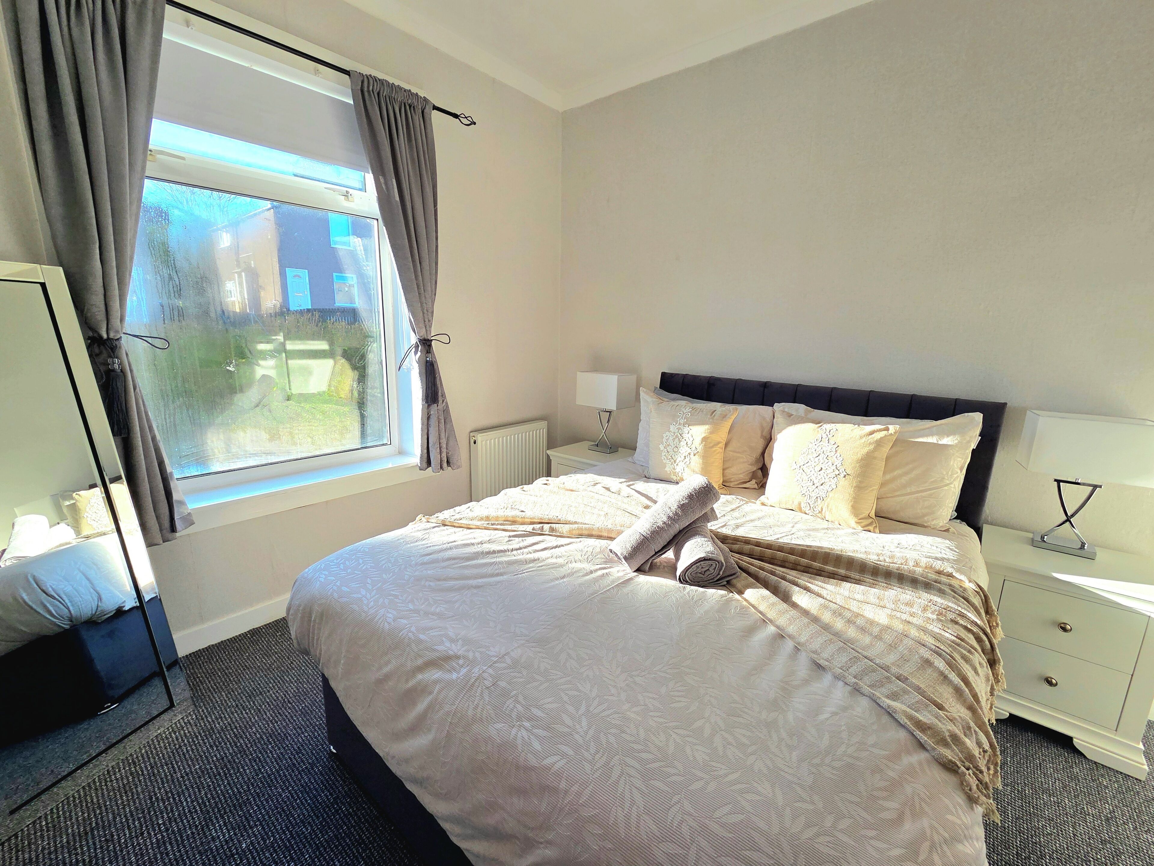 A hotel room with a large bed, a bedside table with a lamp, a window with curtains, and a radiator.