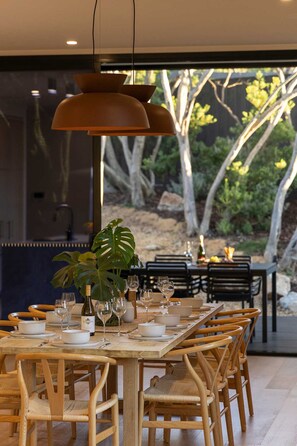 A dining area with a wooden table set for six, surrounded by wicker chairs, with a large potted plant and a hanging pendant light. - The Luxe Seabreeze Heated Pool Spa Cinema (Melbourne)